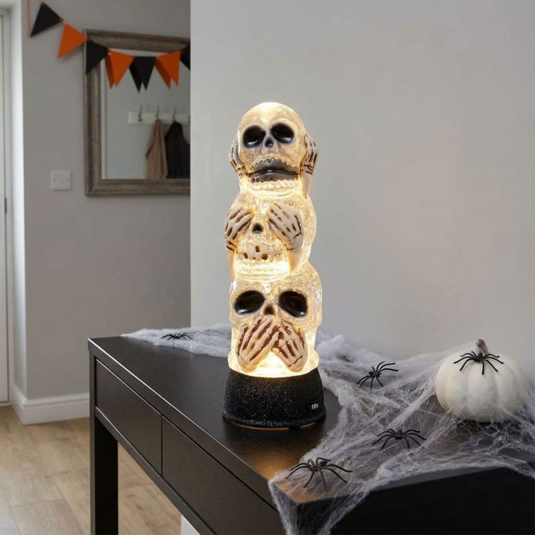A Halloween decorative ornament made of stacked skull figures glowing with warm white light, displayed on a black table with cobwebs, a small pumpkin, and plastic spiders in a home interior.
