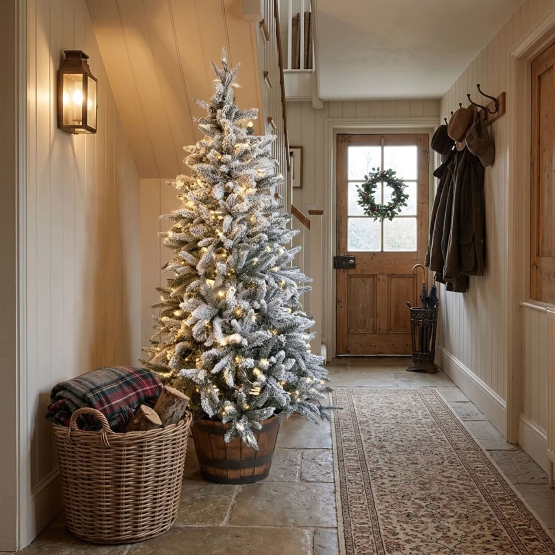 A flocked Christmas tree with warm white string lights in a wooden barrel planter, placed in a cozy hallway next to a wicker basket with a plaid blanket.