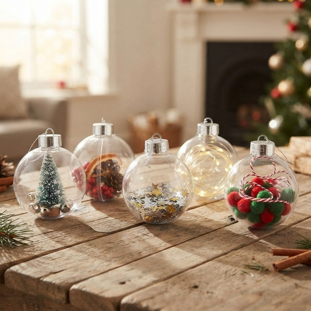 Five clear round Christmas ornaments with silver caps arranged on a rustic wooden table in a cozy living room setting. The ornaments contain festive decorations including a miniature green tree, red berries, gold glitter, and pom-poms.