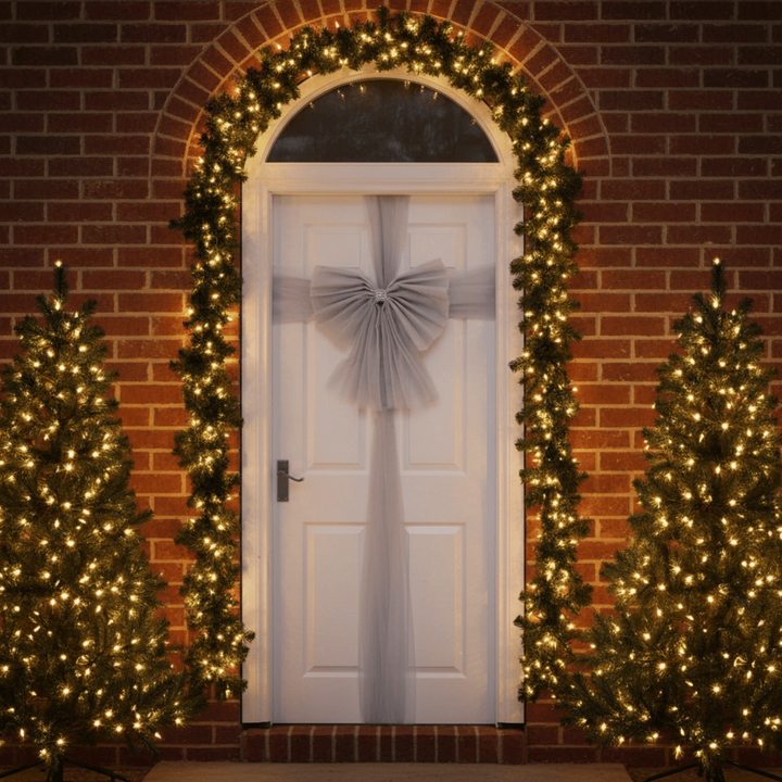 Decorated front door with wreath and bow, flanked by two Christmas trees with lights on a brick wall.