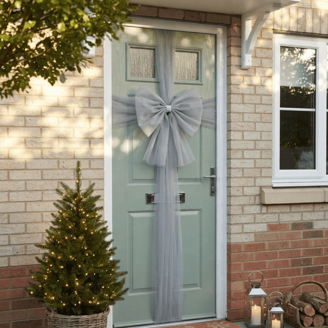 A sage green front door decorated with a large gray tulle bow and matching hanging tulle, shown on a brick house exterior with a small lit Christmas tree and lanterns nearby.