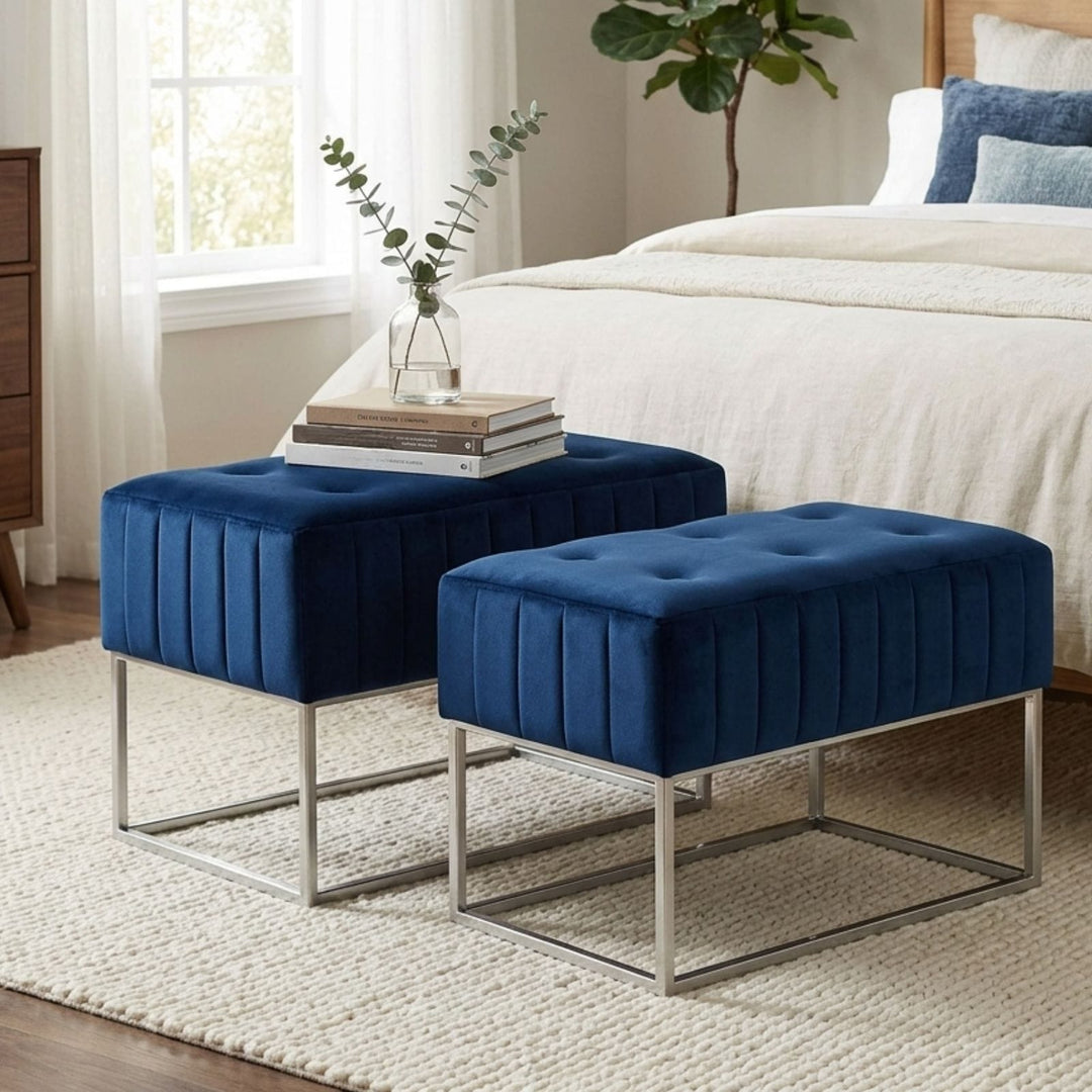 Two navy blue velvet ottomans with silver metal frames in a bedroom setting, styled with books and a vase of eucalyptus on a light beige rug.
