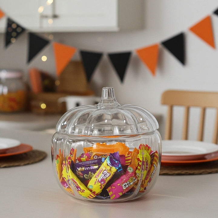 Clear pumpkin shaped candy jar filled with colorful candies on a table with Halloween-themed decorations in the background.