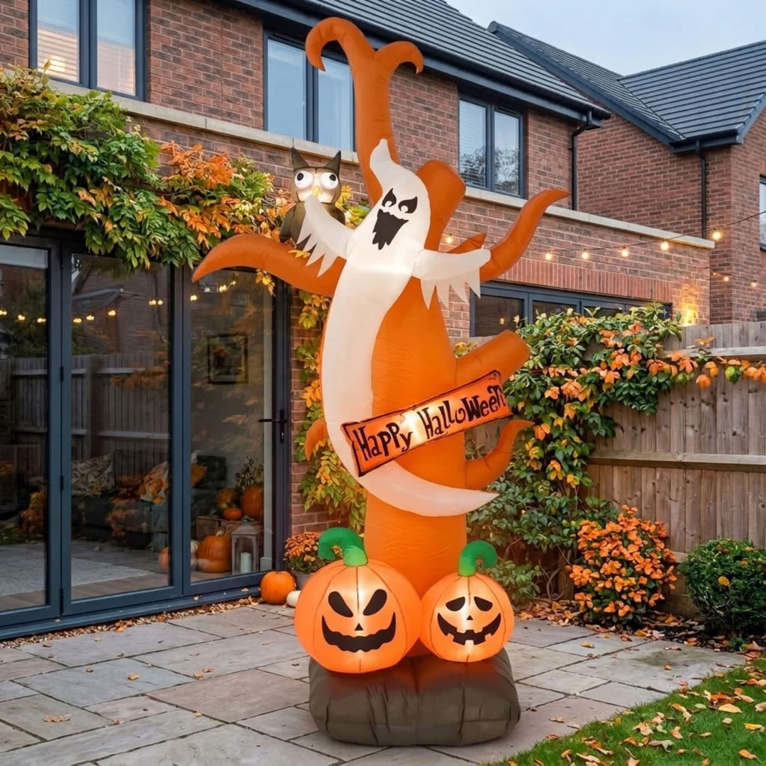 An inflatable Halloween yard decoration stands in a residential backyard. The decoration features a large orange and white ghost holding a 'Happy Halloween' banner, with two glowing jack-o'-lantern pumpkins at its base and a small owl perched on the ghost's shoulder.