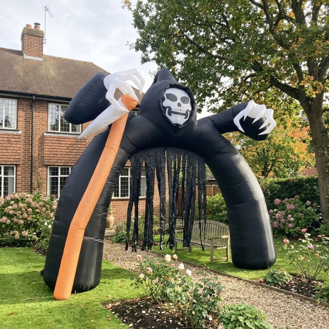 A large inflatable Grim Reaper arch decoration stands on a grassy backyard lawn in front of a brick house. The black inflatable figure has a white skull face, white gloved hands, and holds a large orange scythe.