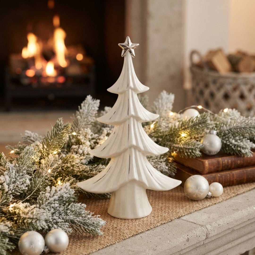 A small white tiered Christmas tree ornament with a silver star topper, displayed on a burlap runner with holiday greenery, silver ornaments, and a warm fireplace in the background.