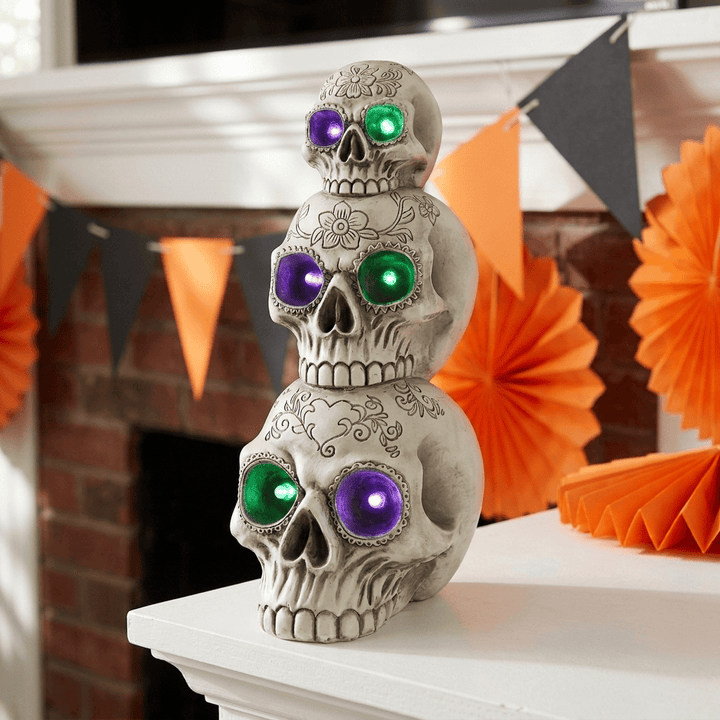 Three stacked skull decorations with glowing multicolor eyes, displayed on a white mantel in front of a brick fireplace with orange paper fans and black-and-orange bunting.