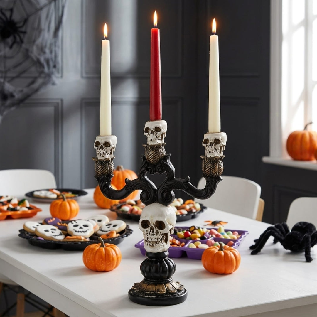 A black metal effect three-arm candelabra centerpiece with skull-shaped candle holders and a central skull base, holding three taper candles (two white, one red), styled on a dining table with Halloween decorations including pumpkins, candy, and a black spider.