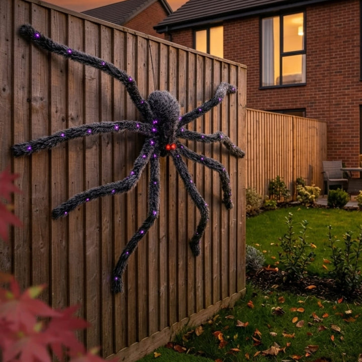 A large Halloween spider decoration mounted on a wooden backyard fence, with glowing purple and red lights along its dark gray legs and body, set against a residential garden at dusk.