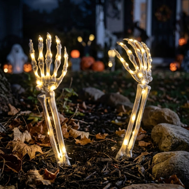 Two skeleton hand Halloween yard lights with warm white LED lights, planted in mulch outdoors at night with a blurred porch and pumpkins in the background.