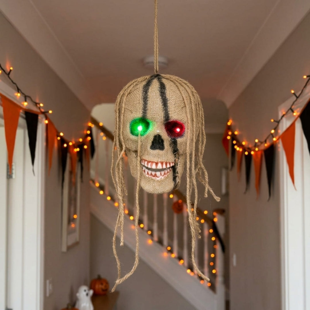A Halloween hanging decoration shaped like a skull with dreadlocks, featuring glowing green and red LED eyes and a toothy grin, suspended in a decorated hallway with orange and black bunting and string lights.