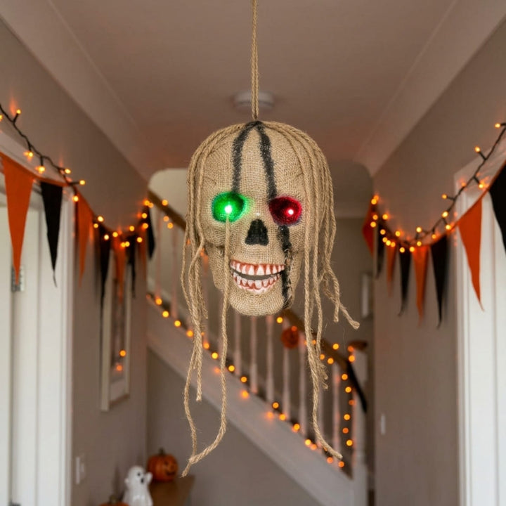 A Halloween hanging decoration shaped like a skull with dreadlocks, featuring glowing green and red LED eyes and a toothy grin, suspended in a decorated hallway with orange and black bunting and string lights.