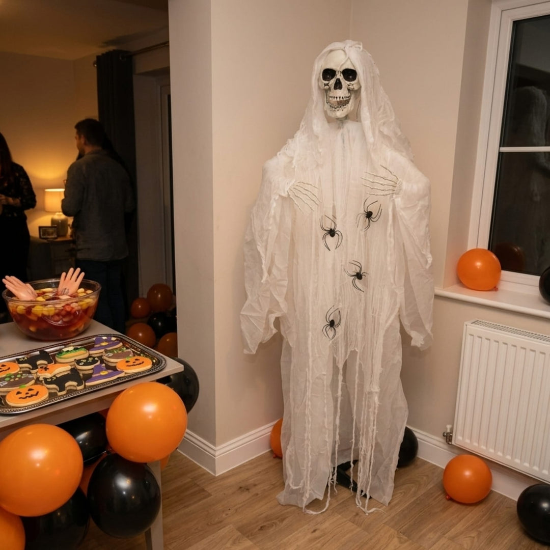A Halloween decoration of a ghostly skeleton draped in white tattered fabric stands in a decorated indoor room with orange and black balloons, a cookie tray, and people visible in the background.