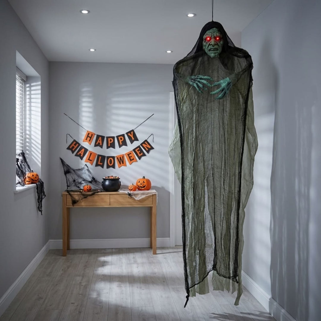 A large hanging Halloween decoration shaped like a ghostly figure draped in tattered black gauze, with a green face and glowing red eyes, displayed in a staged hallway with a 'Happy Halloween' banner and pumpkins on a console table.