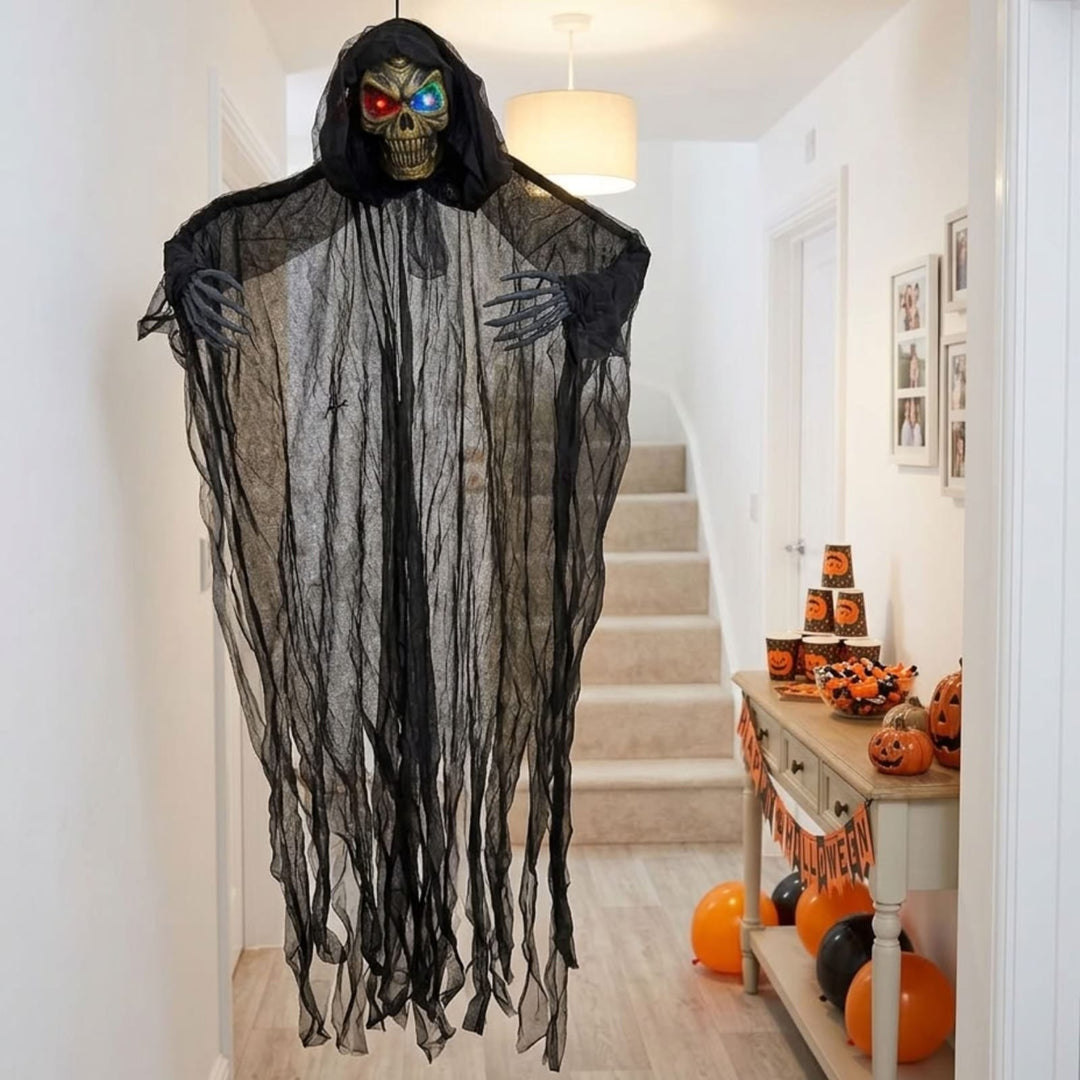 A hanging Halloween decoration shaped like a hooded grim reaper skeleton, with a black tattered robe, gray skeletal hands, and glowing red and blue eyes, displayed in a residential hallway with stairs and a decorated table in the background.