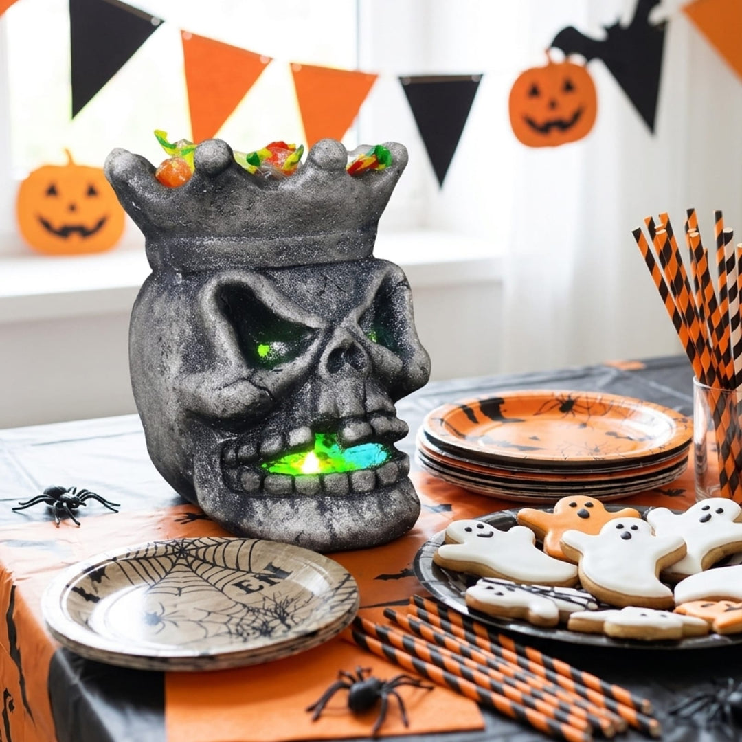 A Halloween-themed tabletop scene with a gray skull-shaped candy holder glowing with multicolor lights, surrounded by orange and black party decorations, plates, cookies, spider props, and striped straws.
