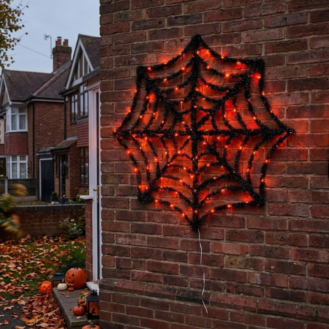 A large black spiderweb-shaped outdoor decoration covered with warm orange lights, mounted on a red brick wall in a residential yard with pumpkins and fallen leaves nearby.