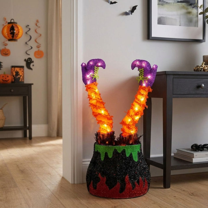 Halloween decoration featuring two glowing orange witch legs with purple feet popping out of a black and red glittery cauldron base with green dripping details, set on a wooden floor in a decorated home interior.