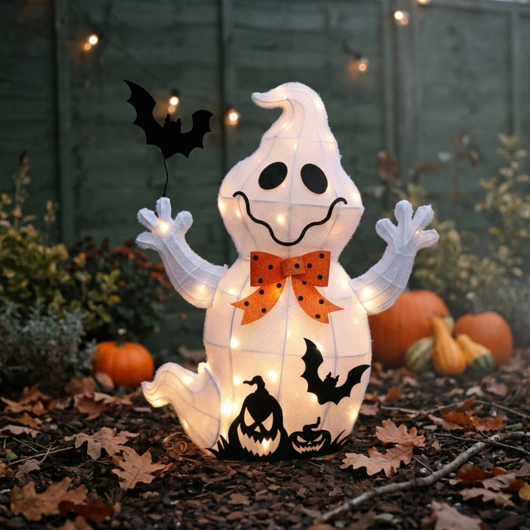 A glowing white ghost-shaped Halloween decoration with black facial features, bat cutouts, and an orange polka-dot bow, displayed outdoors on mulch with pumpkins and string lights in the background.