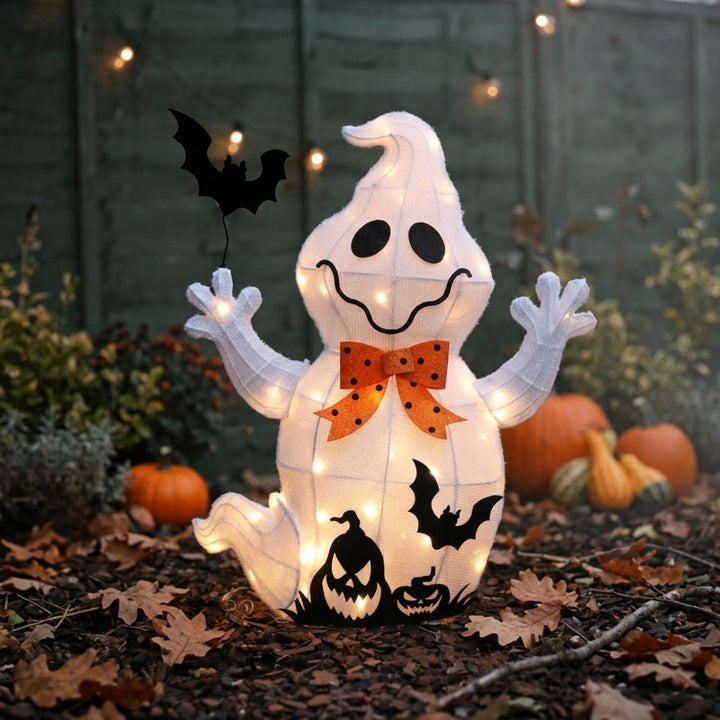 A glowing white ghost-shaped Halloween decoration with black facial features, bat cutouts, and an orange polka-dot bow, displayed outdoors on mulch with pumpkins and string lights in the background.