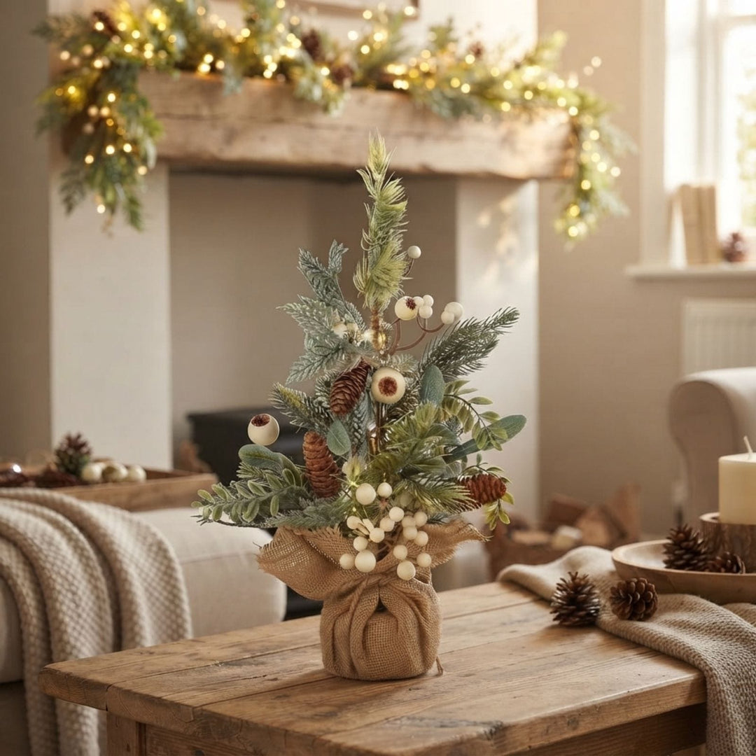 A rustic holiday tabletop arrangement featuring a small artificial Christmas tree with green faux foliage, pinecones, white berries, and a burlap-wrapped base, set on a wooden coffee table in a cosy living room with a fireplace and garland in the background.