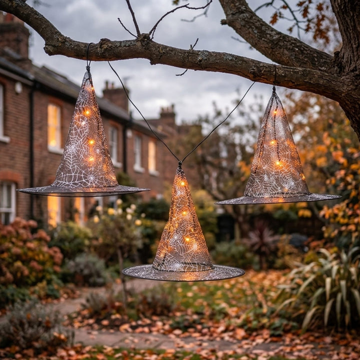 Three witch hat-shaped string lights with spiderweb patterns and warm glowing fairy lights, hanging from a tree branch in a garden setting.