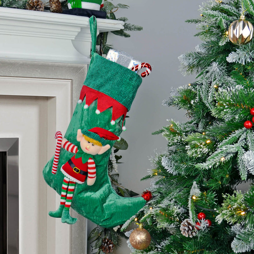Green elf-themed Christmas stocking hanging on a fireplace mantle next to a decorated tree.