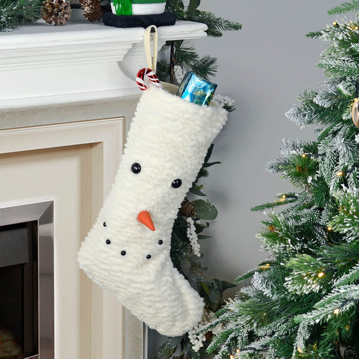Snowman-themed Christmas stocking hanging on a fireplace mantle with a Christmas tree in the foreground.
