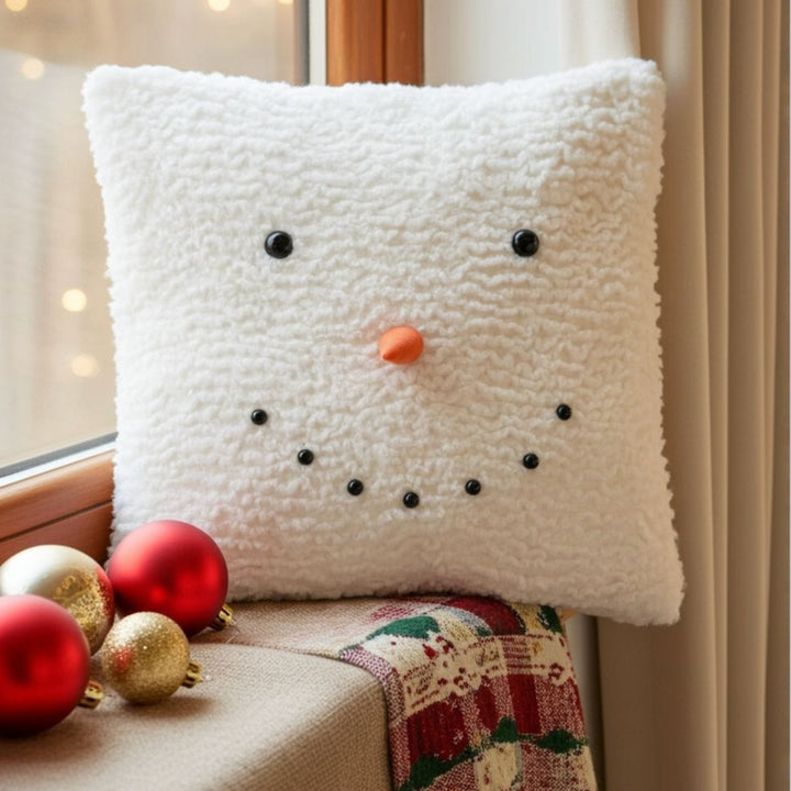 White snowman-shaped cushion with black buttons and an orange carrot nose, placed on a surface with Christmas decorations.