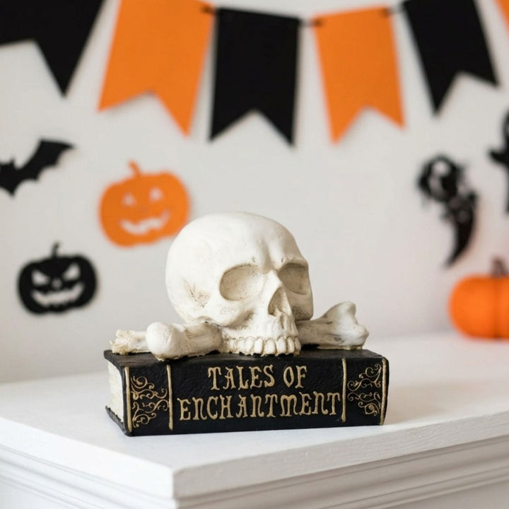 A Halloween decoration featuring a white skull and crossbones resting on a black book with gold lettering that reads 'TALES OF ENCHANTMENT'. The setup is on a white mantel with a blurred background of orange and black Halloween bunting, pumpkins, and a ghost.