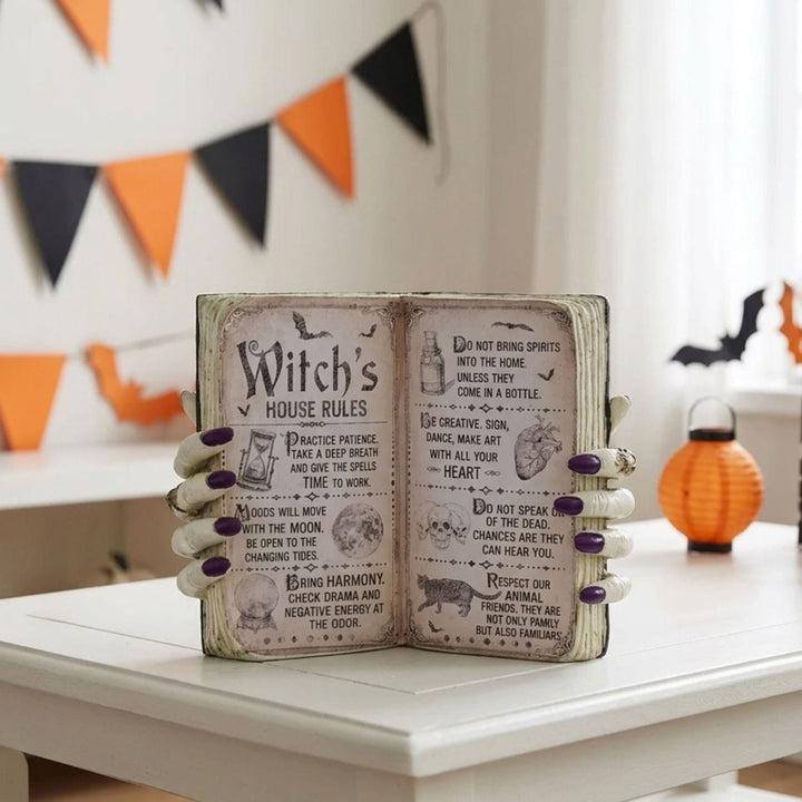 A decorative open book sign with 'Witch's House Rules' printed on the pages, held by two white ghostly hands with purple nail polish. The sign is displayed on a white table with Halloween decorations in the background.