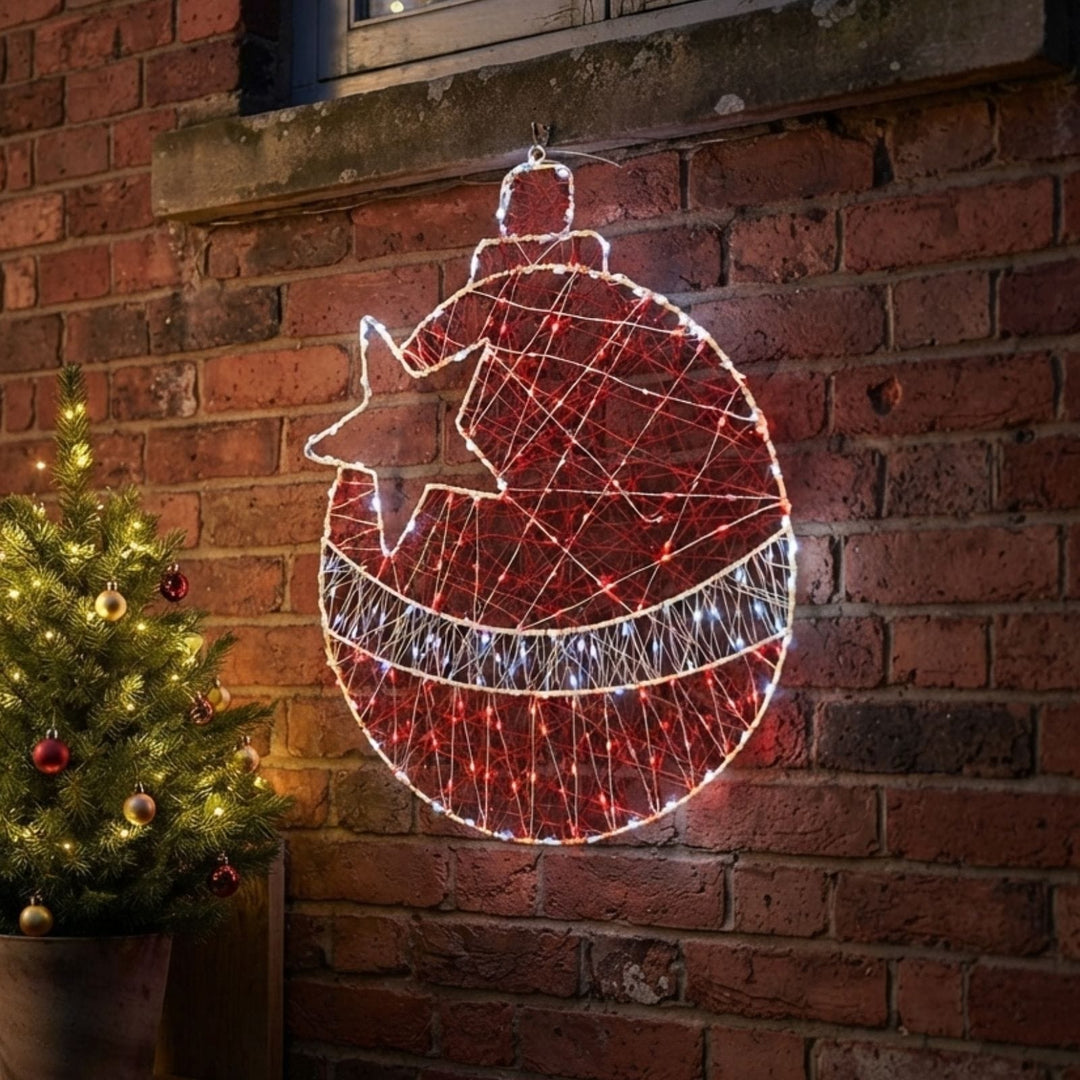A large Christmas ornament-shaped wire light decoration with a star, glowing with red and white lights, hanging on a brick wall next to a lit Christmas tree.