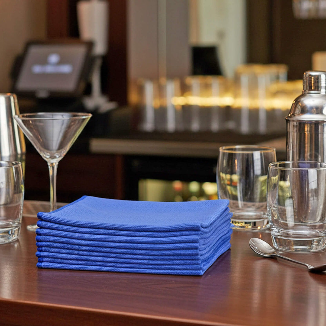 Stack of blue glass cloths on a table with glasses and a cocktail shaker in a blurred bar setting.