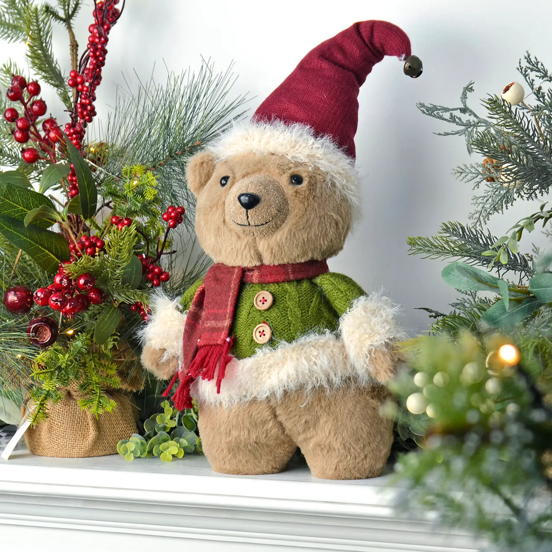 Christmas bear with green jumper and red hat standing on a mantel with tree and lights