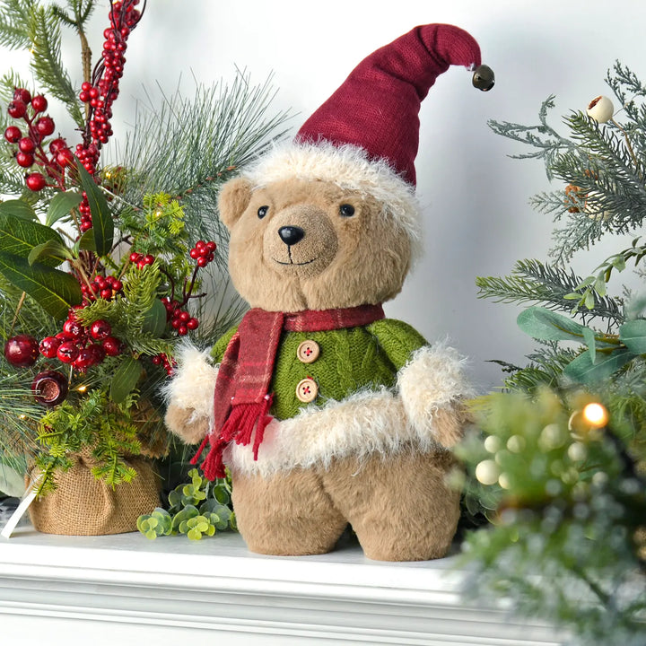Christmas bear with green jumper and red hat standing on a mantel with tree and lights