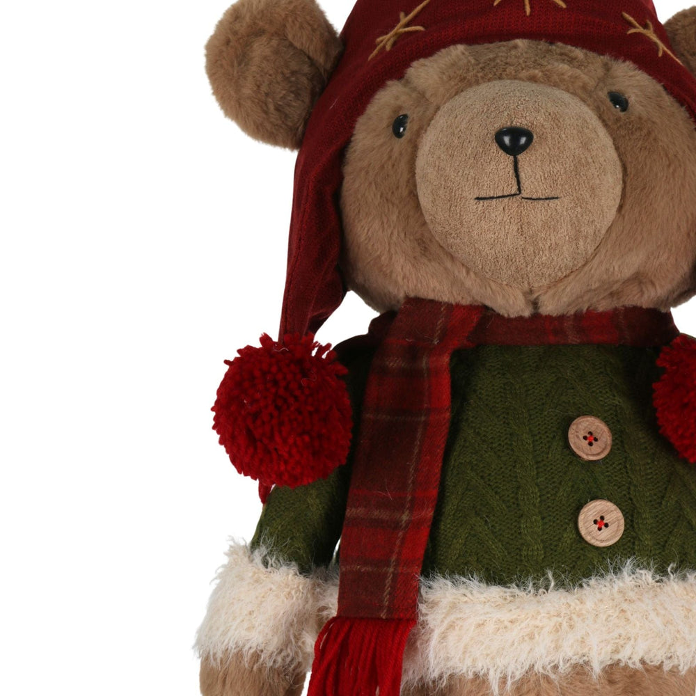 Teddy bear wearing a red hat with pom-poms, green sweater, and red scarf on a white background