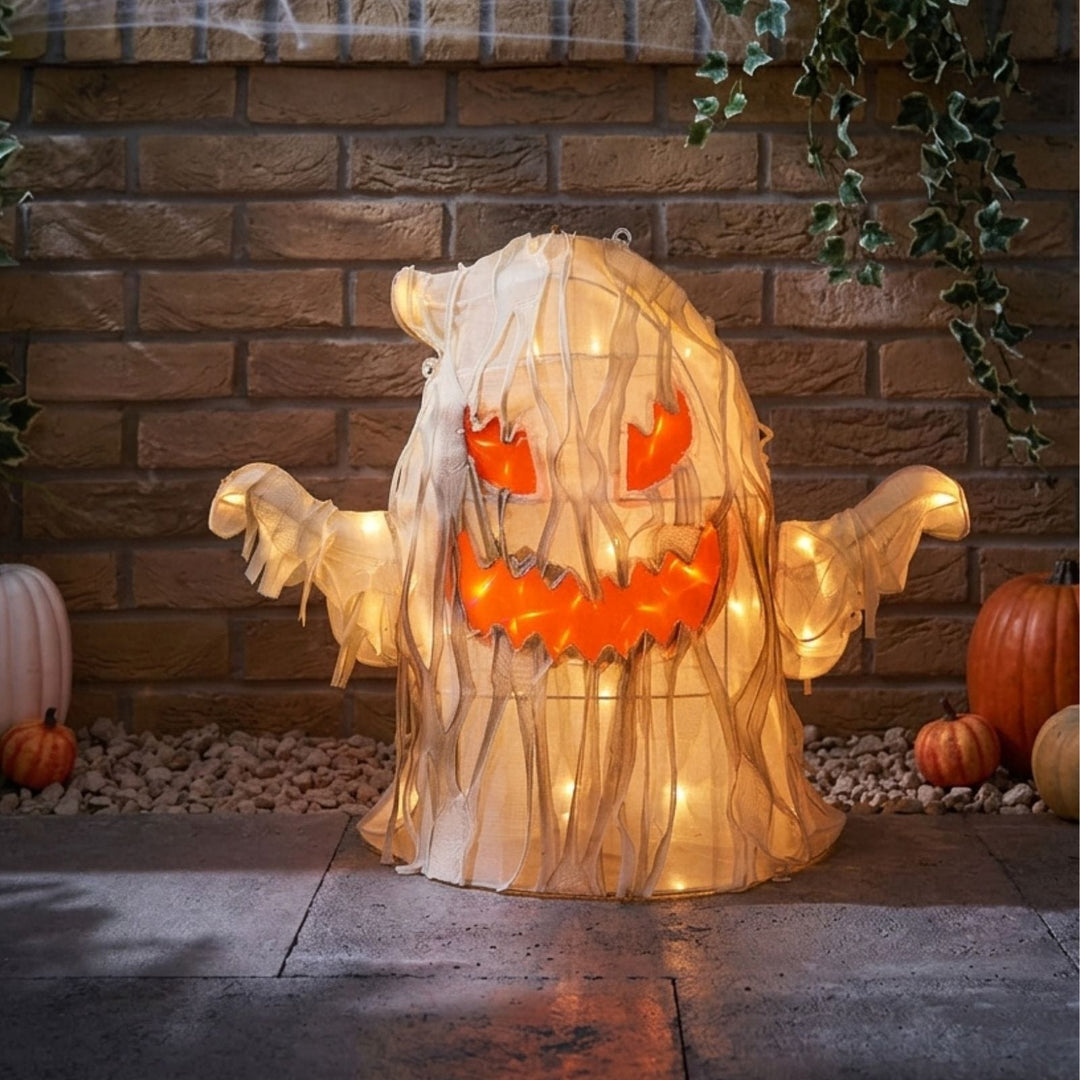 A glowing Halloween ghost decoration draped in translucent white fabric, with orange eyes and a jagged orange mouth, displayed on a stone surface in front of a brick wall with pumpkins and ivy.