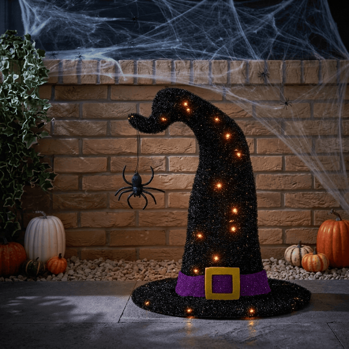 A glittery black witch hat Halloween decoration with a purple band, gold buckle, and warm glowing lights, displayed in a spooky outdoor patio setting with pumpkins, cobwebs, and a spider.
