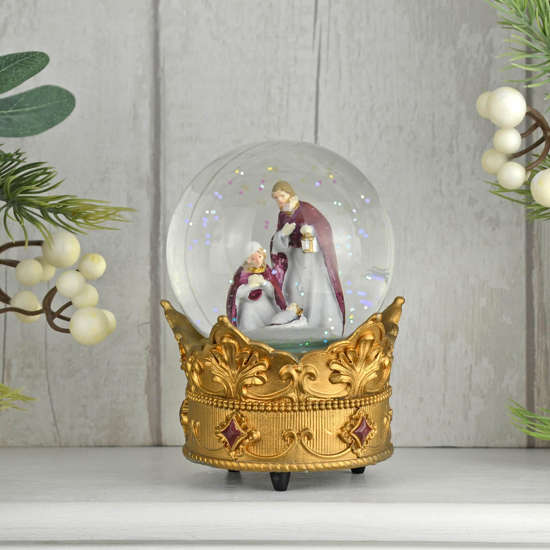 Decorative snow globe with a gold base featuring a Christmas nativity scene, against a light wooden panel background.