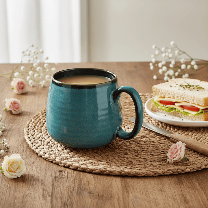 Teal mug with coffee on a wooden table with a sandwich and flowers.