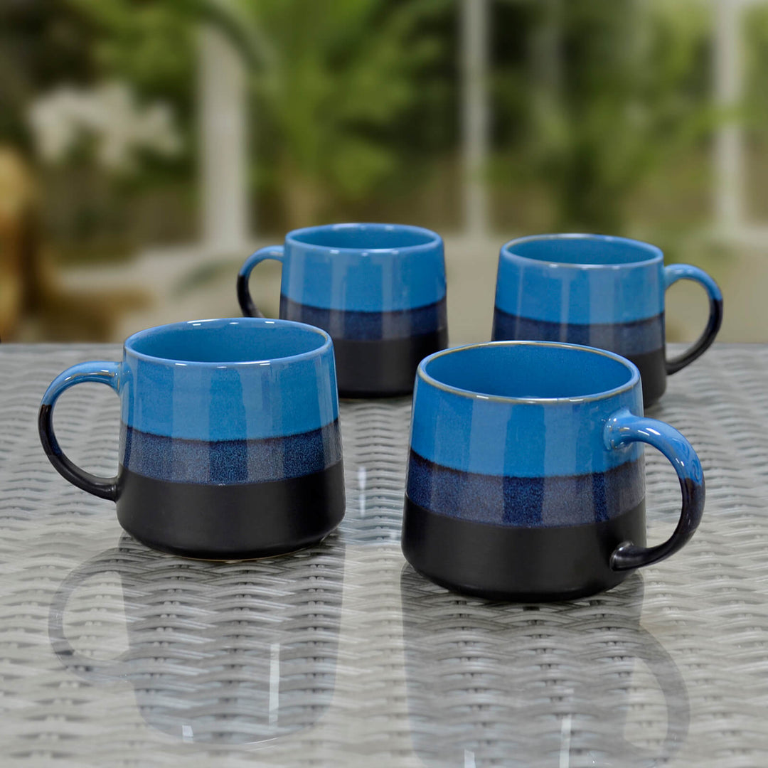 Four blue and black ceramic mugs on a glass topped rattan table with a conservatory background.